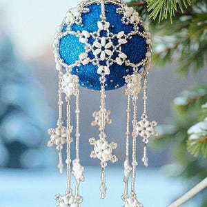 May include: A blue Christmas bauble with white snowflake patterns and dangling beaded embellishments. The bauble hangs from a silver string, with a blurred Christmas tree background.