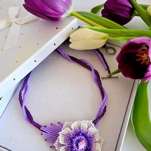 Puede incluir: Un collar de cuentas moradas con un gran colgante de flor. El colgante está hecho de cuentas blancas y moradas y tiene un cristal morado en el centro. El collar está en una caja de regalo blanca con una cinta morada.