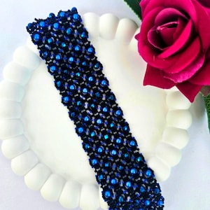 Op de afbeelding: Een handgemaakte armband met een geweven zwart ontwerp met ingebedde blauwe kralen. De armband wordt gepresenteerd op een wit, geschulpte bord, met een levendige rode roos op de achtergrond. Met sluiting.