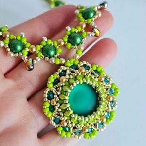 May include: A beaded necklace featuring a large circular pendant with a central matte green cabochon surrounded by intricate layers of lime green, metallic gold, and dark green seed beads, accented with matching beaded chain links.