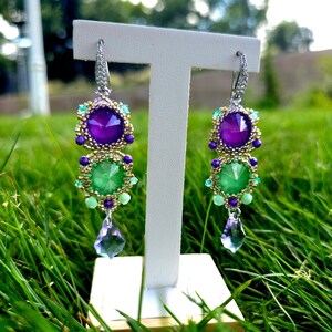Puede incluir: Pendientes colgantes con piedras preciosas moradas y verdes, acentuados con cuentas doradas y turquesas. Los pendientes tienen un cristal en forma de l&aacute;grima en la parte inferior. Se muestran en un soporte blanco.