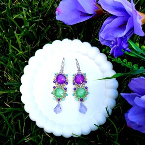 Puede incluir: Un par de pendientes colgantes con un dise&ntilde;o vibrante. Cada pendiente presenta una combinaci&oacute;n de piedras preciosas moradas y verdes, acentuadas con cuentas m&aacute;s peque&ntilde;as. Los pendientes est&aacute;n sobre un plato blanco con bordes festoneados, con flores moradas y hierba verde en el fondo.