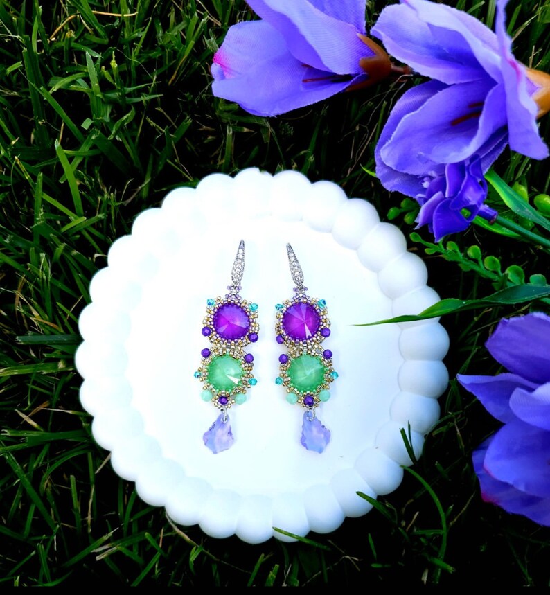 Puede incluir: Un par de pendientes colgantes con un dise&ntilde;o vibrante. Cada pendiente presenta una combinaci&oacute;n de piedras preciosas moradas y verdes, acentuadas con cuentas m&aacute;s peque&ntilde;as. Los pendientes est&aacute;n sobre un plato blanco con bordes festoneados, con flores moradas y hierba verde en el fondo.