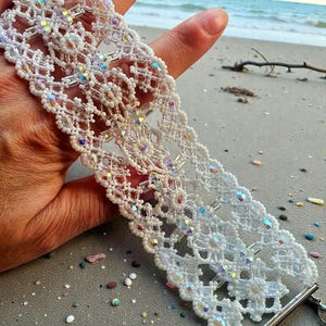 May include: A white beaded bracelet with an intricate openwork design. The bracelet features iridescent crystals and small white beads. The bracelet is held up against a beach background, showcasing its delicate craftsmanship and sparkling details.