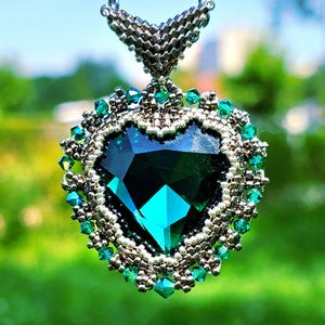 May include: A heart-shaped pendant with a large, faceted emerald-green gemstone. The gemstone is surrounded by a silver-colored beaded frame and smaller green crystals. The pendant hangs from a silver chain, with a blurred green background.