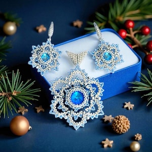 May include: A beaded jewellery set comprising a pendant and matching earrings, each with a snowflake design. The jewellery is mainly light blue and silver, with a large, round, blue centre stone. The set is presented in a blue box, with festive decorations in the background.