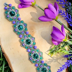 Op de afbeelding: Een kralenarmband met een bloemmotief. De armband heeft smaragdgroene en paarse kralen, met een zilveren sluiting. De armband wordt gepresenteerd op een houten oppervlak, met paarse bloemen en groen in de achtergrond.
