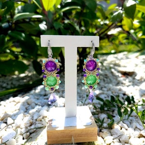 Puede incluir: Pendientes colgantes con un dise&ntilde;o vibrante. Los pendientes presentan una combinaci&oacute;n de piedras preciosas moradas y verdes, acentuadas con cuentas y cristales m&aacute;s peque&ntilde;os. Se exhiben en un soporte blanco con base de madera, sobre un fondo de vegetaci&oacute;n y guijarros blancos.