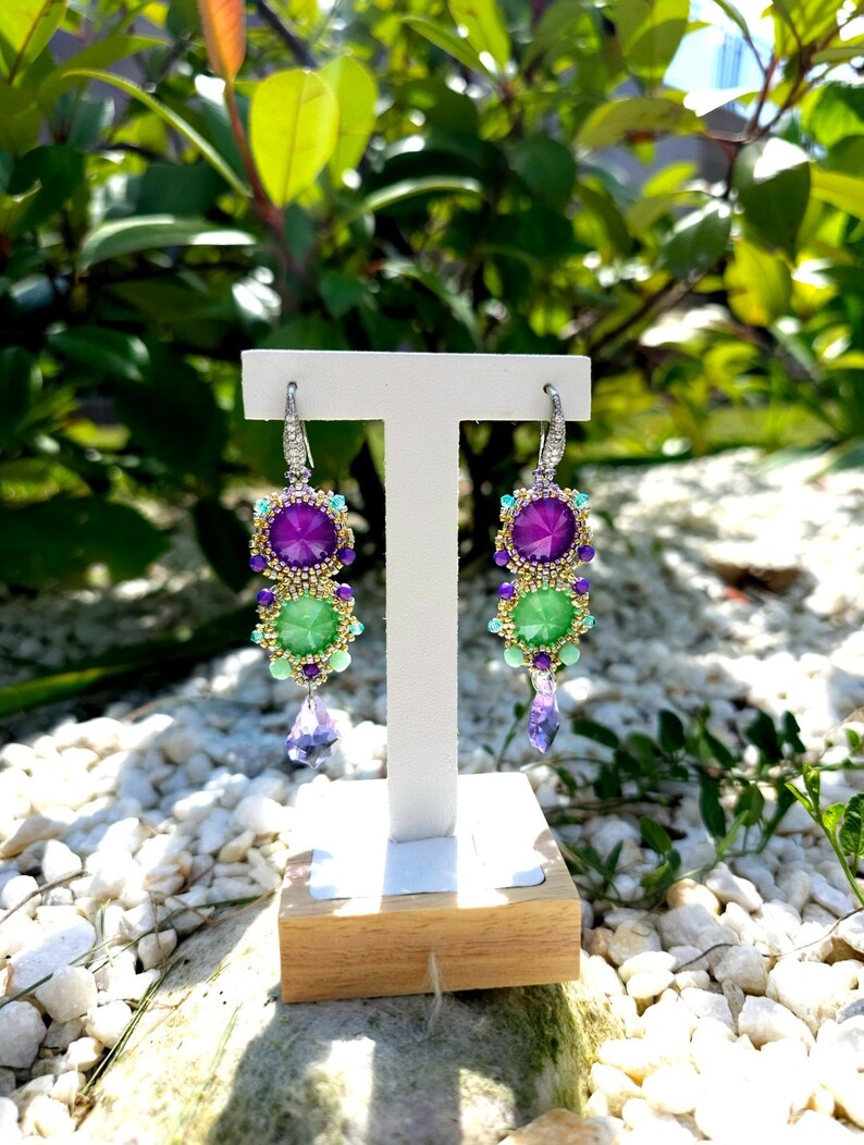 Puede incluir: Pendientes colgantes con un dise&ntilde;o vibrante. Los pendientes presentan una combinaci&oacute;n de piedras preciosas moradas y verdes, acentuadas con cuentas y cristales m&aacute;s peque&ntilde;os. Se exhiben en un soporte blanco con base de madera, sobre un fondo de vegetaci&oacute;n y guijarros blancos.