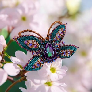 May include: A beaded butterfly brooch with a purple iridescent gemstone in the center. The wings are a mix of teal, purple, and copper beads.