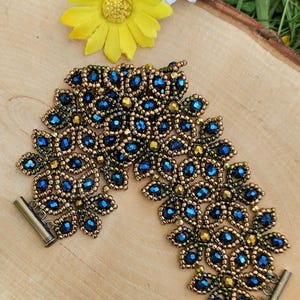 May include: A wide beaded bracelet featuring a repeating floral pattern made of deep blue faceted crystals and metallic gold seed beads, finished with a cylindrical bronze-toned metal clasp at each end of the woven structure.
