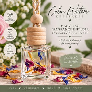May include: A square glass hanging fragrance diffuser bottle filled with dried blue, yellow, and pink flower petals. The bottle features a light wood cap and a braided rope hanger with gold accents. Text reads Calm Waters KEEPSAKES.