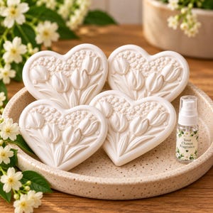 May include: Four heart-shaped white ceramic ornaments featuring embossed tulip and floral patterns. A small clear spray bottle with a label reading Your Chosen Fragrance is positioned next to the ornaments inside a speckled beige circular tray.