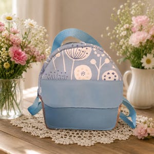 Può includere: Uno zaino azzurro con un motivo floreale e un pannello frontale azzurro chiaro a tinta unita. Lo zaino ha un manico e spallacci blu. Sullo sfondo ci sono vasi di fiori su un tavolo di legno.