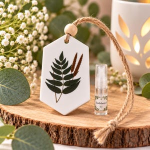 May include: A white hexagonal ceramic hanging ornament featuring a pressed dark green fern leaf and brown grass stalks. The ornament is suspended by a tan jute twine with a wooden bead. A small clear glass spray bottle labelled Sandalwood is included.