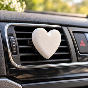 May include: A matte white heart-shaped air freshener clip attached to the horizontal black plastic slats of a vehicle dashboard air vent. The heart is smooth and solid, positioned centrally on the vent, which features a silver trim border.