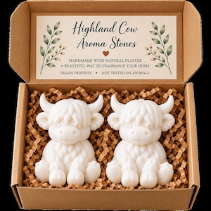 May include: Two white Highland cow-shaped aroma stones made of plaster, nestled in a brown cardboard gift box with crinkle paper. The box lid features the text Highland Cow Aroma Stones, handmade with natural plaster, and vegan friendly.