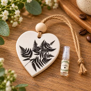 May include: A white ceramic heart-shaped ornament featuring a black fern leaf print, suspended by a tan jute rope with a wooden bead. Beside it sits a small clear spray bottle with a label that reads Coffee and features a small heart icon.