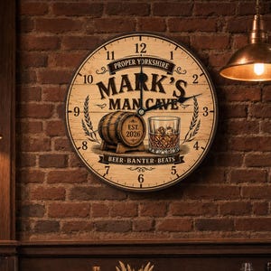 May include: A round wall clock with a light wood grain finish featuring black numerals and hands. The centre displays the text "PROPER YORKSHIRE MARK'S MAN CAVE", an illustration of a wooden barrel, a glass of whiskey, and "EST. 2026" with "BEER BANTER BEATS".