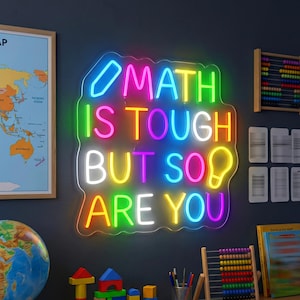 Może przedstawiać: Neonowy szyld zamontowany na ciemnej ścianie z wielokolorowym napisem MATH IS TOUGH BUT SO ARE YOU, zawierający ikonę ołówka w lewym górnym rogu i żarówki w prawym dolnym rogu, podświetlony w żywych odcieniach różu, żółci, zieleni i błękitu.