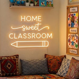 May include: A warm white neon sign mounted on a wall displaying the text HOME sweet CLASSROOM with a pencil graphic underneath. The sign emits a soft glow above a seating area with several patterned throw pillows in various colors and textures.