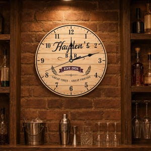 May include: A round wooden wall clock with a light wood grain finish, black numerals, and black hands. The clock face features the text "Hayden's BAR", "EST. 2026", and "GREAT TIMES GREAT FRIENDS" printed in black, with a decorative banner graphic.