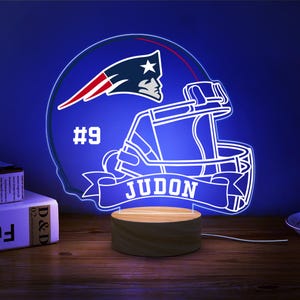 May include: A blue and white football helmet with a white banner that says "JUDON" and the number "#9". The helmet is lit up with blue neon lights and is on a wooden base.