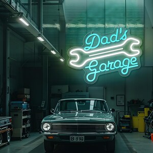 May include: A teal classic car is parked inside a garage. Above the car, a neon sign reads "Dad's Garage" in a script font, with a wrench graphic. The garage has a concrete floor and fluorescent lighting.
