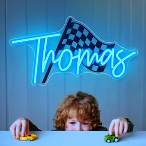 May include: A blue neon sign featuring a chequered flag design with the name "Thomas" in a cursive font. A young child with curly red hair is playing with toy cars below the sign.