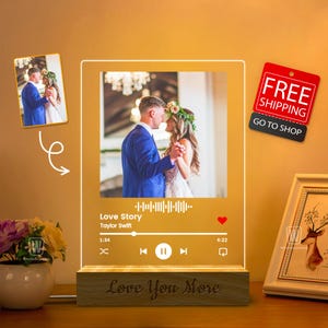 May include: A personalized acrylic photo frame with a built-in music player. The frame displays a wedding photo of a couple dancing. The music player shows the song "Love Story" by Taylor Swift. The base of the frame is wood and has the words "Love You More" engraved on it.