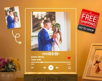 Könnte beinhalten: Ein personalisierter Acryl-Bilderrahmen mit integriertem Musikplayer. Der Rahmen zeigt ein Hochzeitsfoto eines Paares beim Tanzen. Der Musikplayer zeigt den Song "Love Story" von Taylor Swift. Die Basis des Rahmens ist aus Holz und hat die Worte "Love You More" eingraviert.