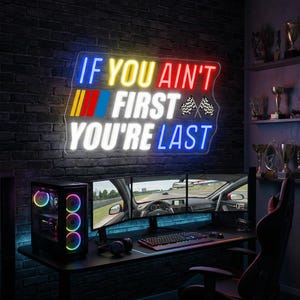 May include: A neon sign with the text "IF YOU AIN'T FIRST YOU'RE LAST" in blue, yellow, red, and white. A gaming setup with a computer, multiple monitors displaying a racing game, and a black desk is below the sign. The background is a brick wall.