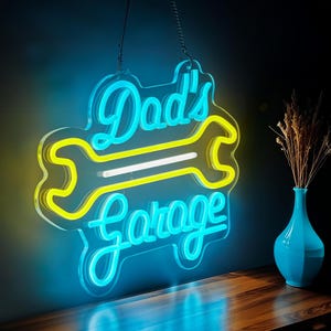 May include: A neon sign with the words "Dad's Garage" in a script font, with a yellow wrench design. The sign is outlined in blue and hangs from a chain. A blue vase with dried flowers sits on a wooden surface.