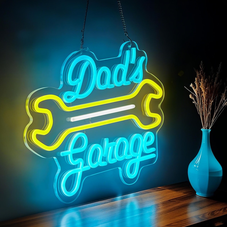 May include: A neon sign with the words "Dad's Garage" in a script font, with a yellow wrench design. The sign is outlined in blue and hangs from a chain. A blue vase with dried flowers sits on a wooden surface.