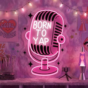 Könnte beinhalten: Ein pinkfarbenes Neon-Mikrofon-Schild mit dem weißen Schriftzug "BORN TO YAP". Das Schild ist an einer pinkfarbenen, flauschigen Wand angebracht, die mit Postern und einer Lavalampe dekoriert ist. Die Szene wird von Lichterketten und einem Make-up-Pinsel-Set beleuchtet.