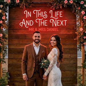 Może przedstawiać: Para pozuje na tle drewnianego tła z neonowym napisem "IN THIS LIFE AND THE NEXT" i "MR & MRS DANIEL". Mężczyzna ma na sobie brązowy garnitur, a kobieta białą koronkową suknię ślubną. Aranżacje kwiatowe otaczają scenę.
