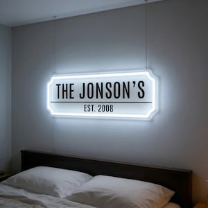 May include: A white neon sign with the text "THE JONSON'S" and "EST. 2008" in black. The sign is rectangular with rounded corners and is hanging above a bed. The sign is illuminated with a bright white light.