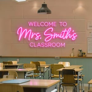 Op de afbeelding: Een felroze neonbord gemonteerd op een lichtgekleurde muur met de tekst WELCOME TO Mrs. Smith's CLASSROOM in een combinatie van blokletters en cursief schrift. Het bord hangt boven een klaslokaal met houten bureaus en stoelen.