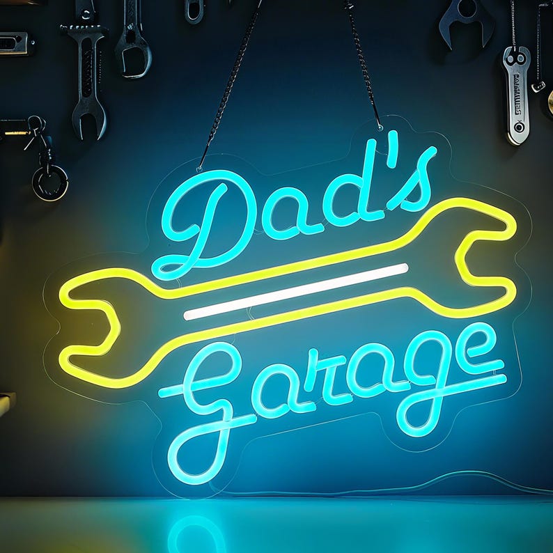 May include: A neon sign with the words "Dad's Garage" in a script font, with a wrench design. The sign has a blue and yellow color scheme and is hanging against a dark background with tools.