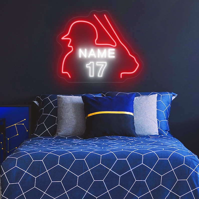Puede incluir: Un letrero de ne&oacute;n rojo que representa a un jugador de b&eacute;isbol con un bate, con el nombre "NAME" y el n&uacute;mero "17" en ne&oacute;n blanco. El letrero est&aacute; montado en una pared azul oscuro sobre una cama azul con ropa de cama de patr&oacute;n geom&eacute;trico blanco.