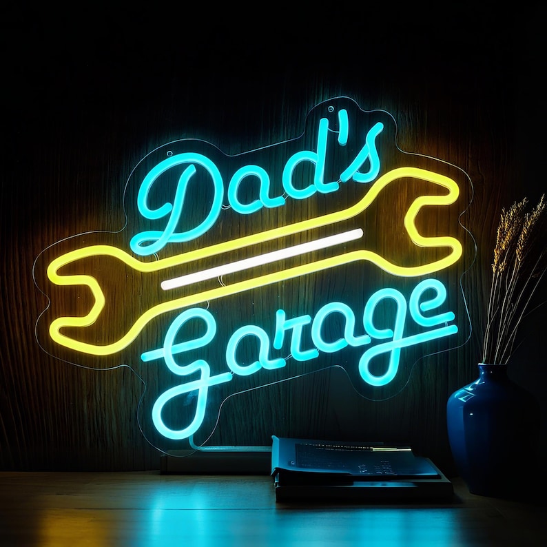 May include: A neon sign with the words "Dad's Garage" in a script font, with a yellow wrench design. The sign has a blue outline and is mounted on a wooden surface. A blue vase with wheat stalks and a stack of books are in the background.