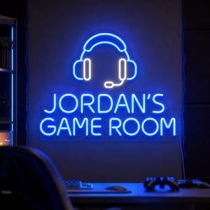 May include: A blue neon sign mounted on a wall featuring a glowing outline of headphones with a microphone above the text JORDAN'S GAME ROOM in a clean sans-serif font. The sign emits a bright blue light against a dark, dim room setting.