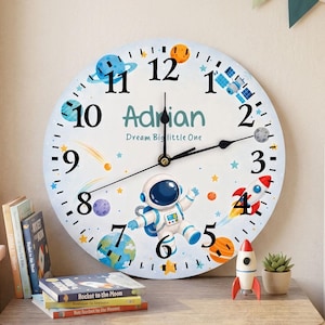 May include: A round wall clock featuring a space theme with an astronaut, planets, stars, and a rocket. The clock face displays the name Adrian and the text Dream Big little One in black numerals and blue lettering with black clock hands.