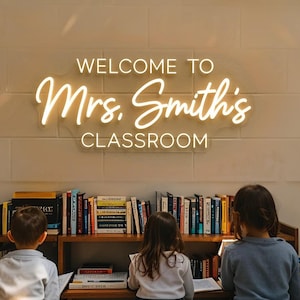 Op de afbeelding: Een aan de muur gemonteerd neonbord met warmwitte verlichting dat de tekst WELCOME TO Mrs. Smith's CLASSROOM weergeeft in een combinatie van blokletters en cursief schrift, geplaatst boven een houten boekenplank vol met diverse boeken.