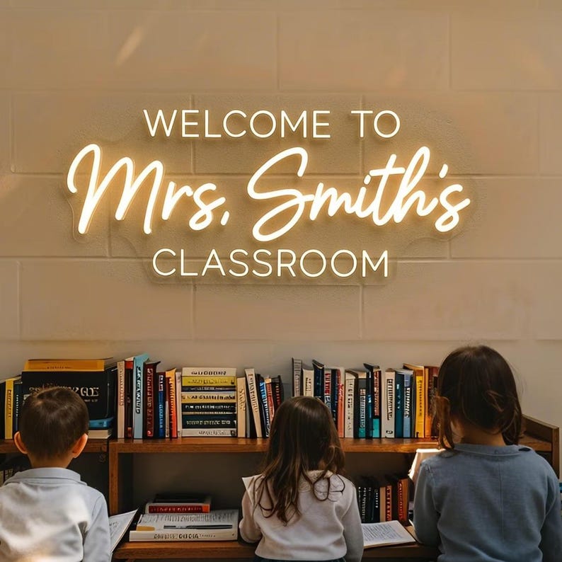 Op de afbeelding: Een aan de muur gemonteerd neonbord met warmwitte verlichting dat de tekst WELCOME TO Mrs. Smith's CLASSROOM weergeeft in een combinatie van blokletters en cursief schrift, geplaatst boven een houten boekenplank vol met diverse boeken.