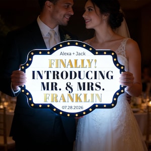 Può includere: Un cartello bianco con scritte dorate e bordo nero, tenuto da una coppia. Il cartello recita "FINALLY! INTRODUCING MR. & MRS. FRANKLIN" con la data 07.28.2026. Il cartello è illuminato da piccole luci.