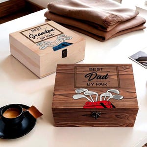 May include: Two wooden boxes with hinged lids. One is light-colored with "Best Grandpa by Par" text and golf club graphics. The other is dark-stained with "Best Dad by Par" text and golf club graphics. A cup of coffee and a blanket are also visible.