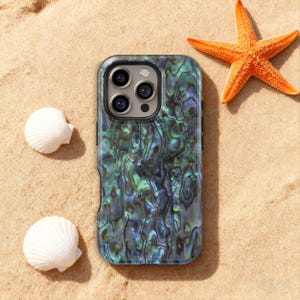 Op de afbeelding: Een smartphonehoesje met een iriserend abalone schelpmotief in groen, blauw en zwart. De hoes ligt op zand, samen met twee witte schelpen en een oranje zeester.