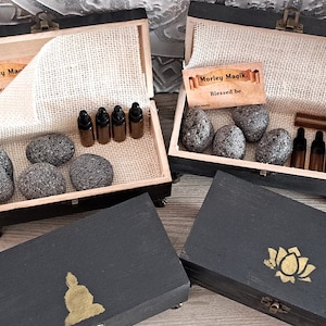 May include: Two wooden boxes with lids open, each containing black stones and small glass bottles. The boxes are decorated with a gold Buddha and a gold lotus flower. The boxes have the text "Morley Magik. Blessed be." on a label inside.