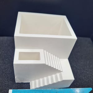 Puede incluir: Una maceta blanca de estilo arquitectónico con una parte superior cuadrada y un diseño de escalera. La maceta tiene una abertura rectangular y una serie de escalones que descienden. La maceta mide aproximadamente 10 cm de ancho.